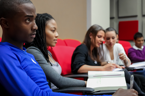 A student in a blue shirt is looking up at a lecturer, pen in hand taking notes. He is surrounded by other note-taking students. 